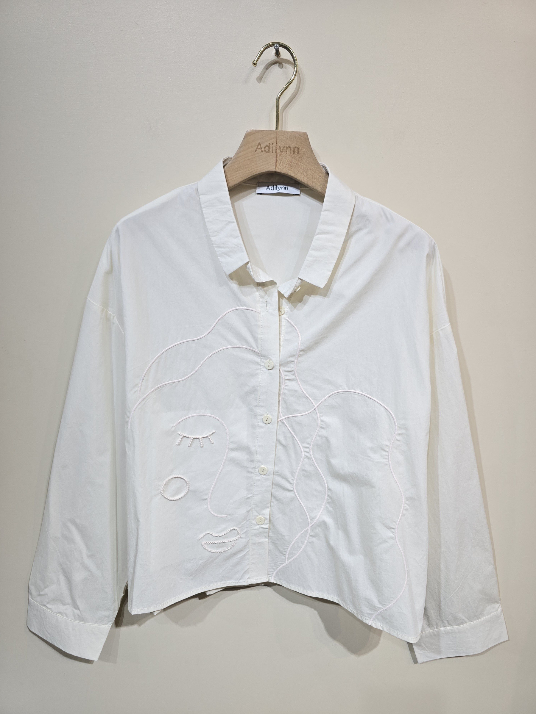 Mayorista Adilynn - Camisa color crudo con botones, cuello clásico, ligeramente holgada y de corte corto. Detalle distintivo: cara bordada en la parte delantera.