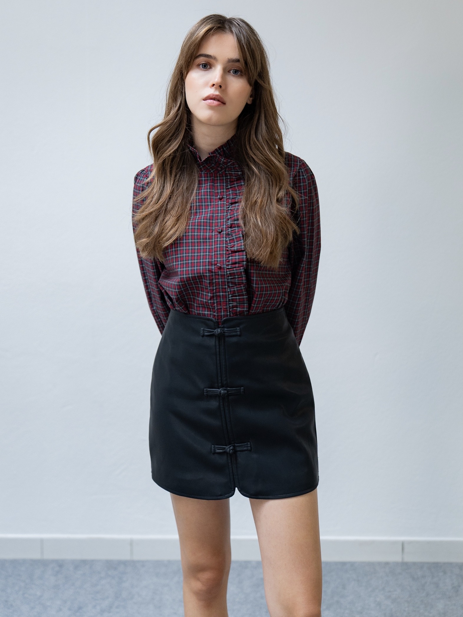 Jupe courte en similicuir.  Elle présente une coupe droite et structurée, animée par des attaches nouées ton sur ton sur le devant, rappelant les détails du style oriental.