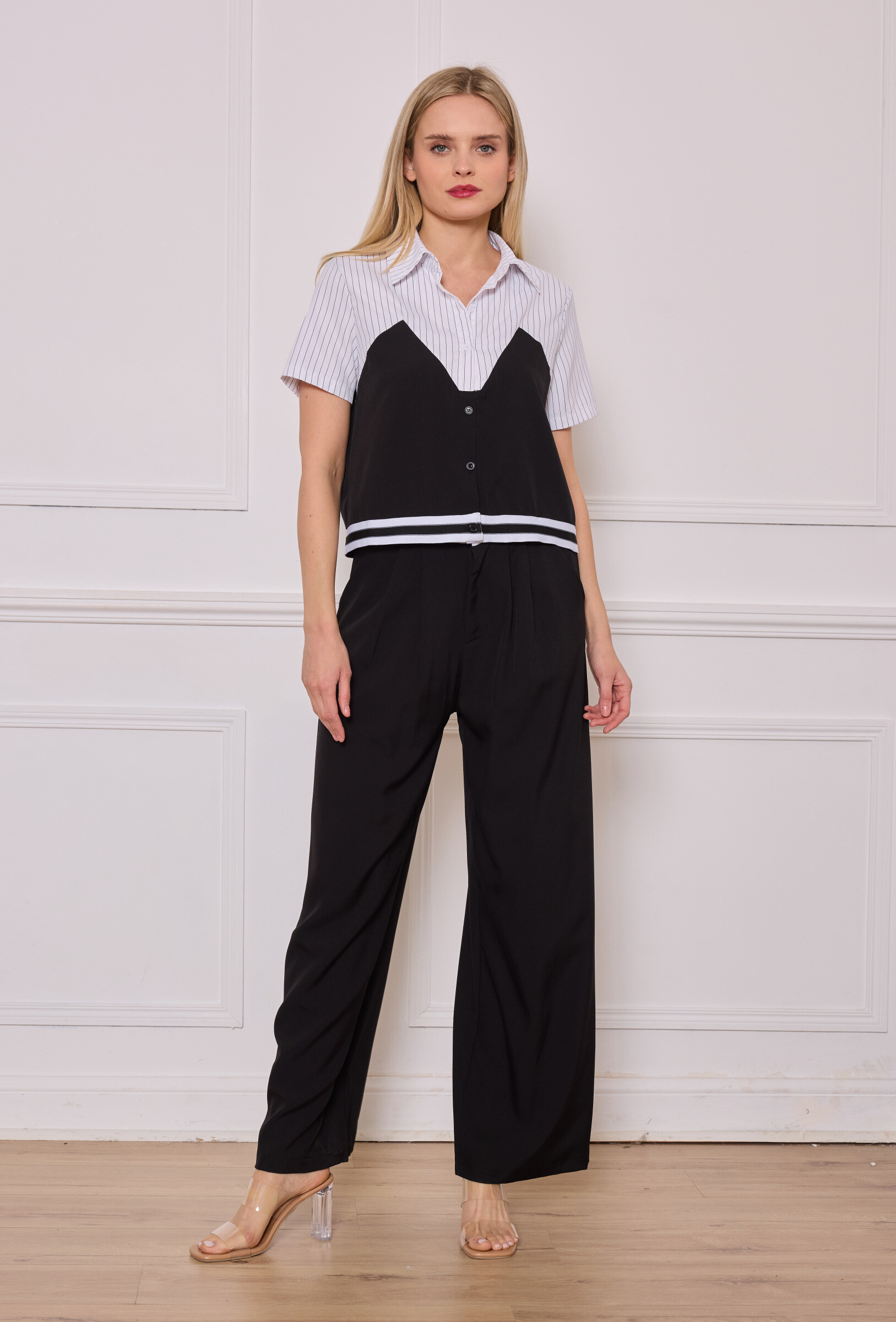 Ensemble deux-pièces au style trompe-l'œil, composé d’un pantalon noir fluide à taille haute et d’un haut effet superposé. Le haut mêle une base de chemise blanche rayée à manches courtes avec un débardeur noir boutonné, souligné par un liseré blanc au bas. Un look structuré et moderne, parfait pour un style casual chic.