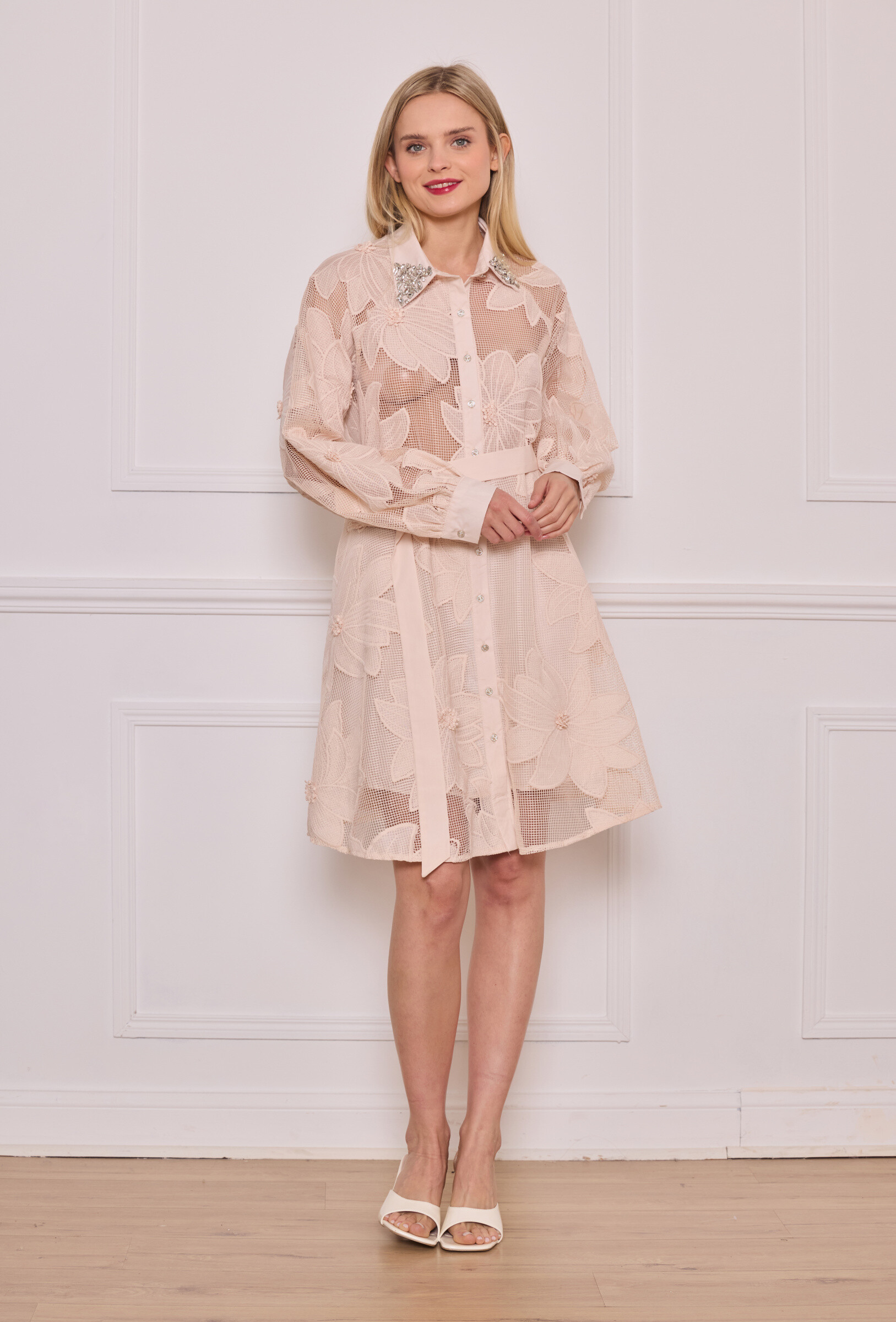 Cette élégante robe chemise en tulle nude séduit par sa broderie florale en relief et sa transparence subtile. Elle est dotée d’un col pointu rehaussé de strass scintillants, d’une fermeture boutonnée sur le devant et d’une ceinture ton sur ton à nouer à la taille pour structurer la silhouette. Une pièce raffinée idéale pour une occasion chic ou une tenue de journée élégante.