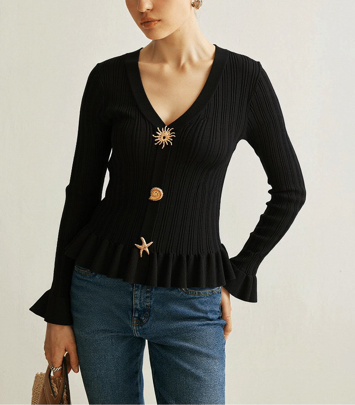 Fine ribbed knit sweater, close to the body, with a V-neckline that highlights the bust line. The long sleeves end with a light ruffle, adding a delicate touch. The bottom of the sweater is also slightly gathered to create a subtle peplum effect. Three decorative buttons — a sun motif, a rounded button and a star — add an original and elegant touch to the center of the room.